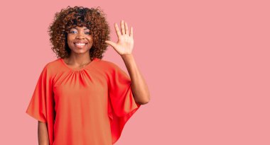 Genç Afro-Amerikalı kadın günlük giysiler giyip beş numaralı parmaklarıyla işaret ederken kendinden emin ve mutlu bir şekilde gülümsüyor.. 