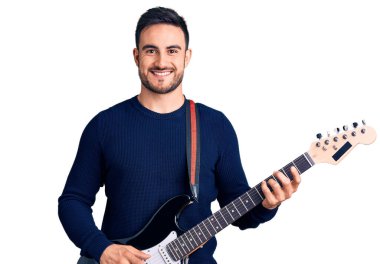 Genç yakışıklı adam elektro gitar çalıyor. Pozitif ve mutlu görünüyor. Kendine güvenen bir gülümsemeyle ayakta duruyor ve dişlerini gösteriyor. 