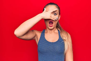 Genç, sarışın, sportif tişörtler giyen, izole edilmiş kırmızı arka planda dikilen, şok halinde yüzünü ve gözlerini kapayarak parmaklarından korkuyla bakan bir kadın.