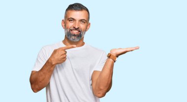 Orta yaşlı, beyaz tişört giyen yakışıklı adam kameraya hayret ediyor ve gülümsüyor.. 