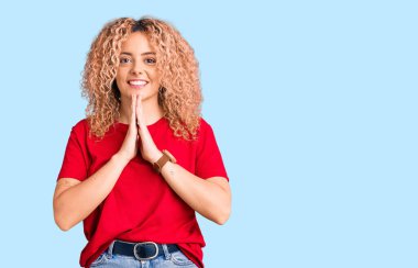 Kıvırcık saçlı, sıradan kırmızı tişörtlü, elleri birarada dua eden, affedilmek isteyen, kendinden emin gülümseyen genç sarışın kadın.. 