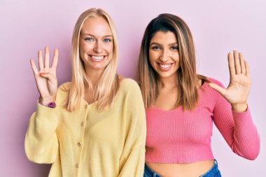 Pembe arka planda yan yana duran iki arkadaş dokuz numaralı parmaklarıyla işaret ederken kendinden emin ve mutlu bir şekilde gülümsüyor.. 
