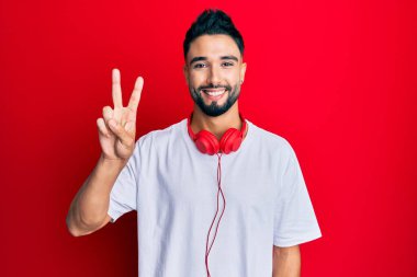 Sakallı genç adam kulaklık kullanarak müzik dinliyor. İki numaralı parmaklarıyla işaret ediyor. Kendinden emin ve mutlu bir şekilde gülümsüyor.. 