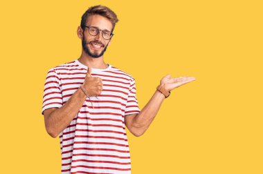 Sakallı, sıradan kıyafetler ve gözlük takan yakışıklı sarışın adam avuçlarını gösteriyor ve baş parmaklarıyla hareket ediyor, mutlu ve neşeli gülümsüyor. 