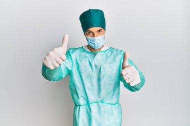 Cerrah üniforması giyen ve tıbbi maske takan genç bir adam elleriyle olumlu bir jest yapmayı onaylıyor, gülümsüyor ve başarı için seviniyor. kazanan jest. 