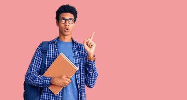 Afrika kökenli Amerikalı genç bir adam sırt çantası takıyor. Şaşırmış ve şaşırmış bir şekilde yukarı bakıyor ve parmaklarını gösterip kollarını kaldırıyor.. 