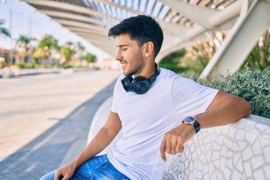 Genç Latin adam mutlu bir şekilde müzik dinliyor. Parktaki bankta oturmuş kulaklık takıyor..