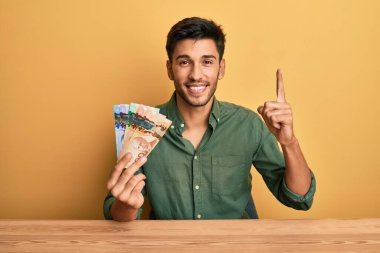 Kanada doları tutan genç yakışıklı adam şaşırmış, şaşırmış ve parmaklarıyla işaret ediyor ve kollarını kaldırıyor. 