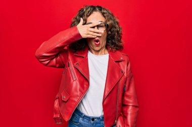 Orta yaşlı, güzel bir kadın, kırmızı ceket ve gözlük takıyor, izole edilmiş bir arka planda, şok halinde yüzünü ve gözlerini eliyle kapatıyor, parmaklarından korkuyla bakıyor.
