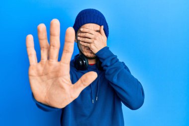 Eşofman altı ve kulaklık takan İspanyol genç adam elleriyle gözlerini kapatıyor ve üzgün ve korku ifadeleriyle jestleri durduruyor. utangaç ve olumsuz kavram. 