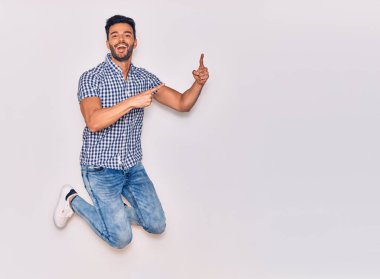 Genç, yakışıklı, sıradan kıyafetler giyen, mutlu gülümseyen bir İspanyol. Yüzünde tebessümle sıçrayıp, izole beyaz arka planda parmaklarını yana doğru gösteriyor.