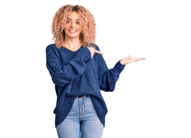 Kış süveteri giyen kıvırcık saçlı genç sarışın kadın kameraya hayret ediyor ve gülümsüyor.. 