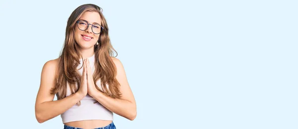 カジュアルな服と眼鏡を手で祈っている美しい白人の若い女性が一緒に自信を笑顔許しを求めて — ストック写真