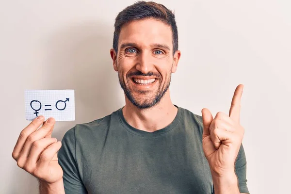 Young handsome man asking for sex discrimination holding paper with gender equality message smiling happy pointing with hand and finger to the side