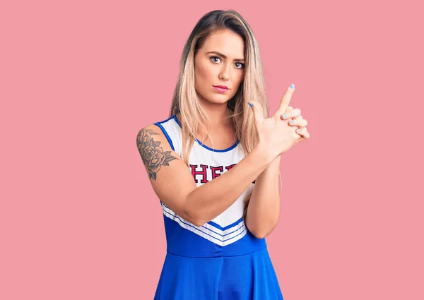 Young beautiful blonde woman wearing cheerleader uniform holding symbolic gun with hand gesture, playing killing shooting weapons, angry face