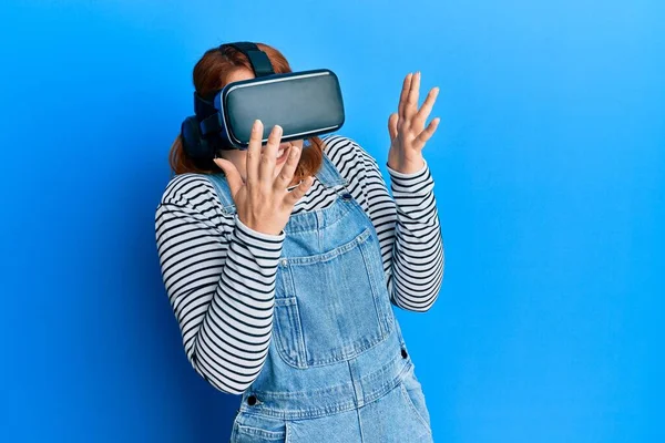 Young redhead woman scared using 3d virtual glasses standing over isolated blue background.