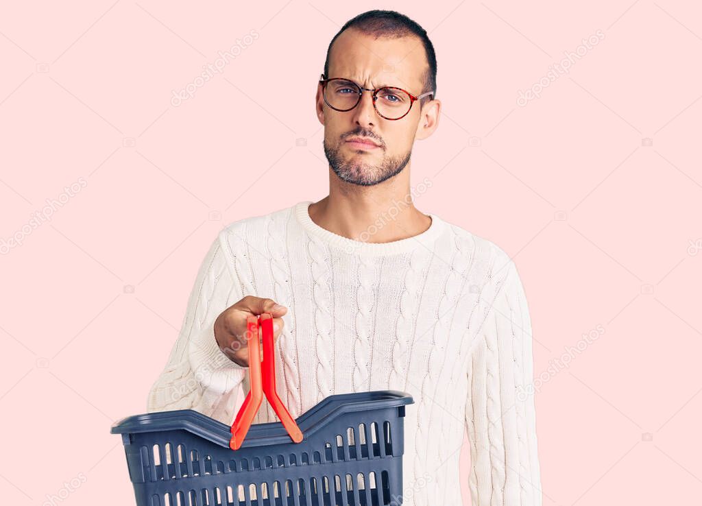 Young handsome man holding supermarket shopping basket thinking attitude and sober expression looking self confident 