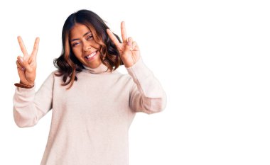 Balıkçı yaka kazak giyen, ağzı açık gülümseyen ve parmaklarını iki eliyle zafer işareti yapan güzel, melez bir kadın. İki numara.. 