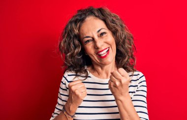 Orta yaşlı, esmer, çizgili tişört giyen güzel bir kadın kırmızı arka planda dikilmiş şaşırmış ve şaşırmış bir şekilde kolları havada ve gözleri kapalı bir şekilde başarıyı kutluyor.