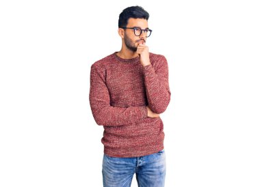 Kışlık süveter giyen ve eli çenesinde gözlük takan genç, yakışıklı İspanyol adam soru sormayı düşünüyor, düşünceli bir ifade. Düşünceli bir yüzle gülümsüyor. şüphe kavramı. 