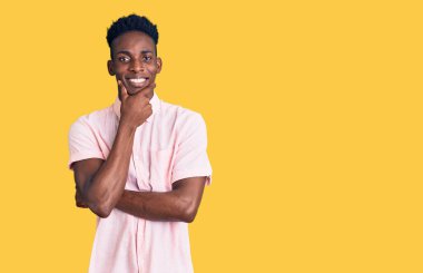 Sıradan kıyafetler giyen genç Afro-Amerikalı adam kameraya kendinden emin bakıyor kollarını kavuşturmuş ve çenesini kaldırmış gülümsüyor. Olumlu düşünmek. 