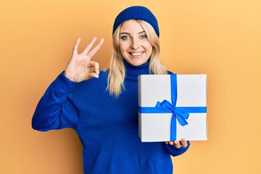 Beyaz bir kadın elinde hediye tutuyor. Parmaklarıyla imza atıyor. Gülümsüyor. 