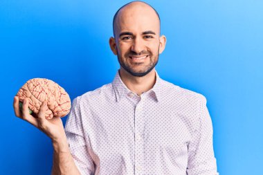 Beyni tutan genç yakışıklı kel adam pozitif ve mutlu görünüyor. Kendine güvenen bir gülümsemeyle ayakta duruyor ve dişlerini gösteriyor. 