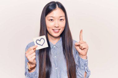 Genç, güzel Çinli bir kadın kalp şeklinde hatırlatıcı taşıyor. Bir fikir veya soruyla gülümsüyor. Mutlu bir yüzle işaret ediyor. 