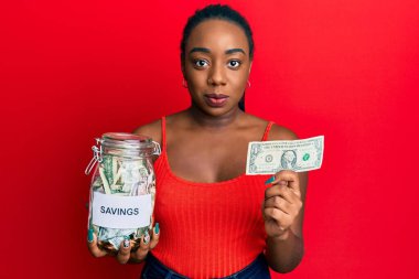 Genç Afro-Amerikalı bir kadın elinde kavanozla 1 dolarlık banknot tutuyor ve yüzünde ciddi bir ifadeyle rahatlıyor. Basit ve doğal bir şekilde kameraya bakıyorsun.. 