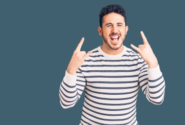 Latin asıllı genç adam, gündelik kıyafetler giyip çılgın bir ifadeyle bağırıyor ve elleri havada rock sembolü yapıyor. Müzik yıldızı. ağır müzik konsepti. 