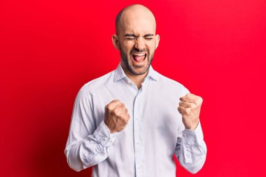 Zarif bir gömlek giyen yakışıklı kel adam şaşkınlık ve şaşkınlık içinde kollarını kaldırarak ve gözlerini kapayarak başarıyı kutluyor. 