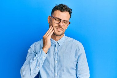 Günlük kıyafetler giyen ve gözlüklü genç bir İspanyol diş ağrısı ya da diş hastalığından dolayı ağızlarına acı dolu bir ifadeyle dokunuyor. dişçi 