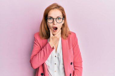 İş stili ve gözlük takan genç beyaz bir kadın inançsızlığa, şaşkınlığa ve elleri çenesinde şaşkınlığa kapılmış bir ifade takınıyor. 