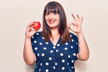 Genç ve cüsseli bir kadın kırmızı elma tutuyor. Parmaklarıyla imza atıyor. Gülümsüyor. 