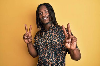 Saçları örgülü, leopar desenli hayvan desenli tişörtlü, parmaklarını zafer işareti yaparken gösteren kameraya bakan Afro-Amerikan bir adam. İki numara.. 