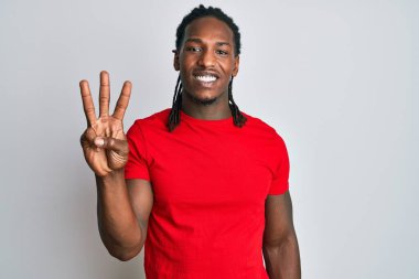 Saçları örgülü Afro-Amerikalı bir adam üç numarayı gösterip gösterirken kendinden emin ve mutlu gülümsüyor.. 