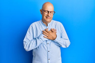 Orta yaşlı kel bir adam günlük kıyafetler ve gözlük takıyor elleri göğsünde, gözleri kapalı gülümsüyor ve yüzünde minnet dolu bir jest var. sağlık kavramı. 