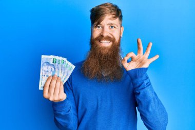 Genç İrlandalı kızıl saçlı adam Güney Afrika 'yı elinde tutuyor. 100 rand banknot. Tamam işareti yapıyor. 