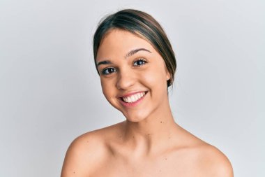 Genç esmer kadın üstsüz duruyor. Cildi pozitifmiş ve mutlu görünüyor. Kendine güvenen bir gülümsemeyle dişlerini gösteriyor. 