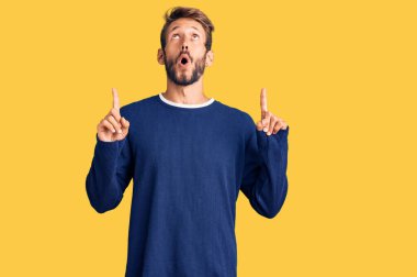 Sarışın, sakallı, sıradan bir süveter giyen yakışıklı adam şaşırmış ve şaşırmış bir şekilde yukarı bakıyor ve parmaklarıyla işaret ediyor ve kollarını kaldırıyor.. 