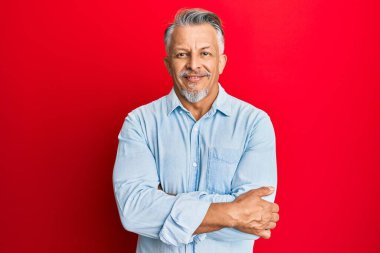 Orta yaşlı, gri saçlı, sıradan kıyafetler giyen mutlu yüzlü, kollarını kavuşturup kameraya bakan bir adam. pozitif kişi. 