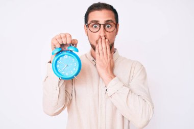Çalar saati tutan genç yakışıklı adam ağızlarını eliyle kapatıyor, şok olmuş ve hata yapmaktan korkuyor. şaşırmış ifade 