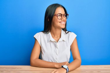 Güzel İspanyol kadın, gündelik giysiler içinde masada oturuyor, yüzünde gülümsemeyle yan yana bakıyor, doğal bir ifade. Kendinden emin gülen. 