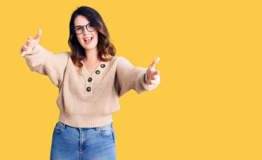 Güzel, genç, esmer bir kadın, günlük kıyafetler ve gözlük takmış, kameraya bakıyor ve sarılmak için kollarını açmış gülümsüyor. Mutluluğu kucaklayan neşeli bir ifade.. 