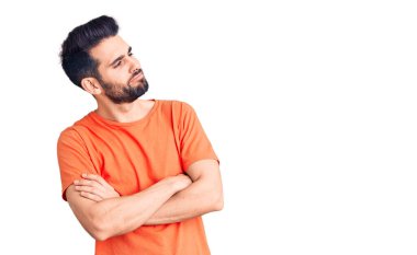 Sakallı, sıradan tişörtlü, kolları çapraz ve kendinden emin genç bir adam. 