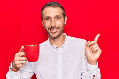 Genç yakışıklı adam elinde kahveyle gülümsüyor mutlu bir şekilde elini ve parmağını yana doğru gösteriyor. 