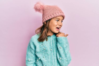 Yün süveter giyen güzel bir kız ve sevimli bir şapkayla baş parmağını yana uzatarak gülümsüyor. 