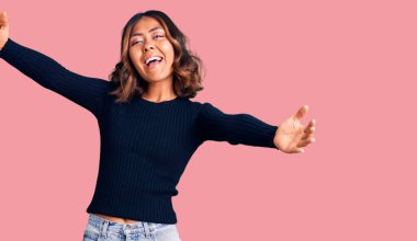 Genç, güzel, melez bir kadın, günlük giysiler içinde kameraya bakıyor ve sarılmak için kollarını açmış gülümsüyor. Mutluluğu kucaklayan neşeli bir ifade.. 