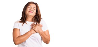 Orta yaşlı, beyaz tişört giyen, göğsünde elleri kapalı, yüzünde minnet ifadesi olan Latin bir kadın. sağlık kavramı. 