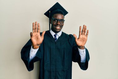 Mezuniyet şapkası ve tören cübbesi giyen yakışıklı siyah adam dokuz numarayı gösterip işaret ederken kendinden emin ve mutlu bir şekilde gülümsüyor.. 